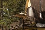 A rustic A-frame cabin with a wooden deck and umbrella sits among tall pine trees. The cabin has a sloped roof and a chimney, with a gravel driveway leading up to it. The surrounding area is lush with greenery and dappled sunlight.