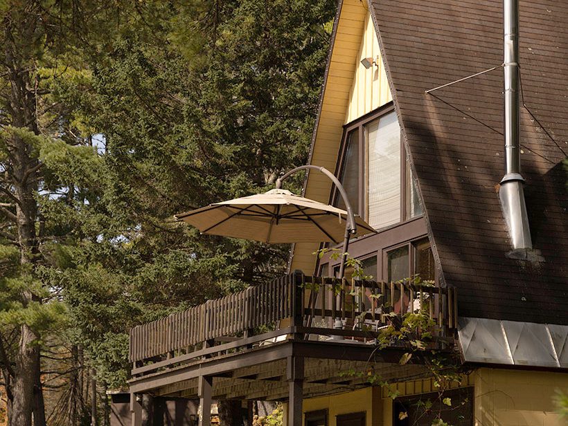 A rustic A-frame cabin with a wooden deck and umbrella sits among tall pine trees. The cabin has a sloped roof and a chimney, with a gravel driveway leading up to it. The surrounding area is lush with greenery and dappled sunlight.