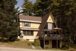 A two-story A-frame house with a sloped roof and wooden exterior sits surrounded by tall trees. The house features multiple windows, a balcony with a table and umbrella, a garage on the lower level, and satellite dishes attached to the side.