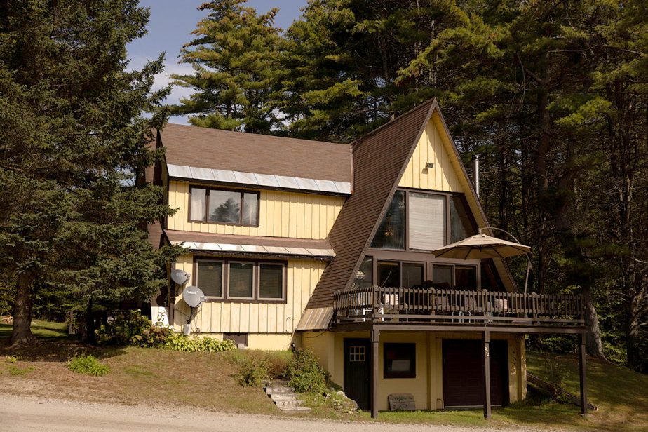 A two-story A-frame house with a sloped roof and wooden exterior sits surrounded by tall trees. The house features multiple windows, a balcony with a table and umbrella, a garage on the lower level, and satellite dishes attached to the side.