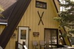 A cozy A-frame cabin with wide yellow wooden panels, brown trim, and a peaked roof. The porch features two chairs, a small table, and flower pots, while a pair of skis is mounted above the window. Trees surround the cabin, adding to the rustic charm.