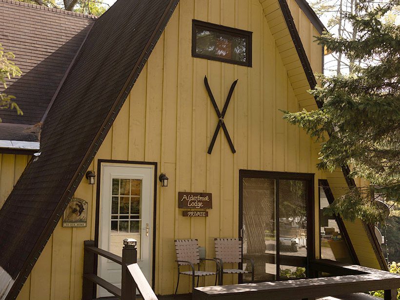 A cozy A-frame cabin with wide yellow wooden panels, brown trim, and a peaked roof. The porch features two chairs, a small table, and flower pots, while a pair of skis is mounted above the window. Trees surround the cabin, adding to the rustic charm.