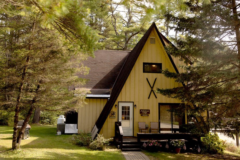 A cozy A-frame cabin surrounded by lush trees. The house features yellow paneling, a dark brown triangular roof and decorative crossed skis on its front. A small deck with chairs and potted plants is at the entrance. Bright daylight filters through the trees.