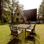 A backyard featuring a large glass-top table surrounded by eight metal chairs on a grassy lawn. The house in the background has an A-frame section with yellow siding and tall trees providing a forest-like environment. Sunlight filters through the trees.