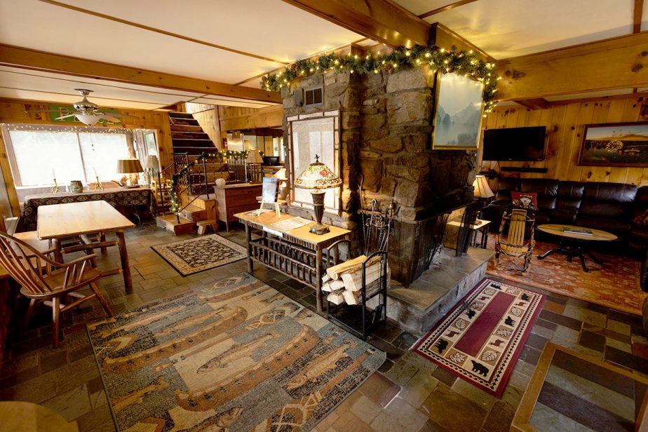 A cozy rustic living room featuring stone fireplace adorned with string lights, wooden furniture with patterned cushions, dining table, area rugs, and stairs leading to second floor. Warm lighting enhances the wood-paneled walls and assorted decor.