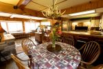 A cozy kitchen and dining area with wooden furniture and walls. A round table covered with a patterned tablecloth featuring moose and trees has a vase with flowers. An antler chandelier hangs above. Windows in the background let in natural light.