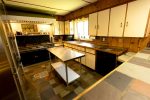 A warm, rustic kitchen with wood-paneled walls, white cabinets, and black countertops. A stainless steel island sits in the center, while a large window over the sink lets in natural light. The kitchen has a traditional stove and tiled flooring with a mix of colors.