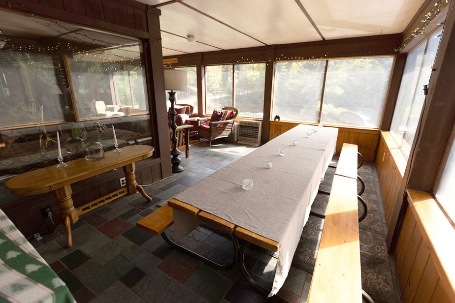 A sunlit dining room with large windows features a long table covered in a beige cloth, accompanied by wooden benches. The room includes a wooden side table with decorative bottles and cozy sitting area with two red armchairs and a small table near the windows.