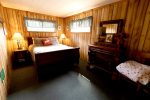 A cozy wooden cabin bedroom with wood paneling on the walls, a bed with decorative pillows and a duvet, and a nightstand with a lamp. A dresser with a mirror, a vase of flowers, and framed artwork is on the right. Two windows with blue curtains let in natural light.