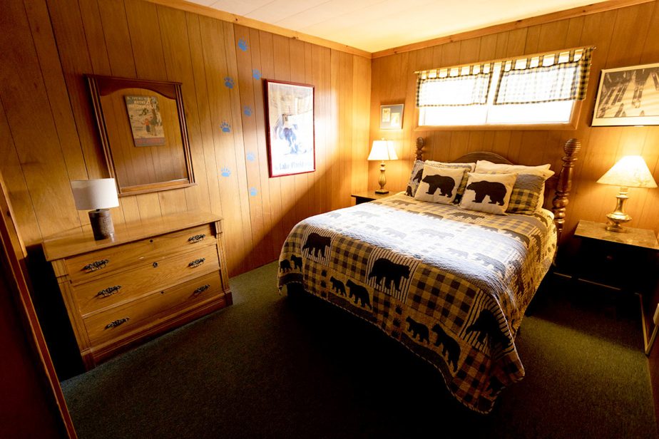 A cozy cabin bedroom with wood-paneled walls features a bed adorned with a checkered quilt displaying bear silhouettes. The room includes a wooden dresser, bedside tables with lit lamps, artwork on the walls, and a window with a plaid valance.