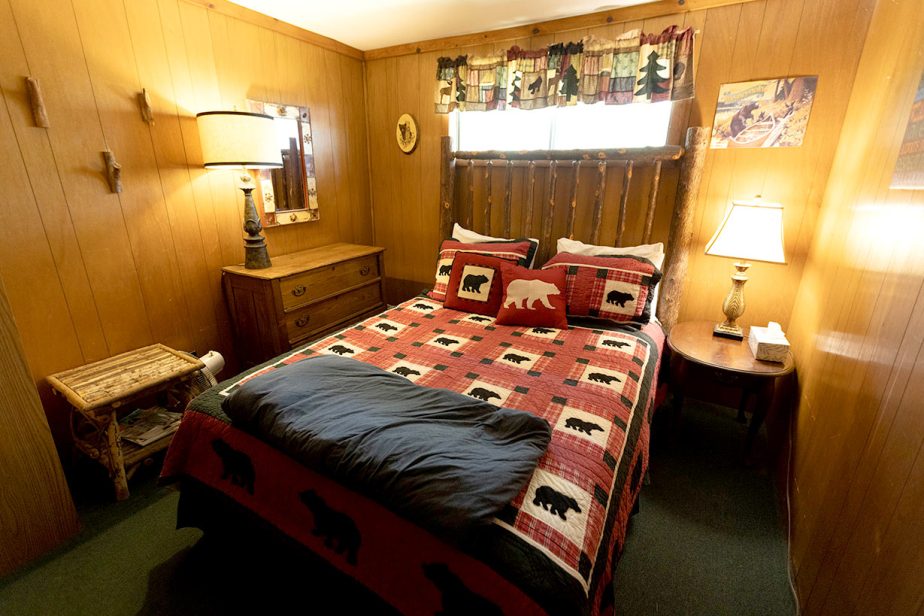 A cozy log cabin bedroom with wood-paneled walls features a bed with a rustic wooden headboard, adorned with a red and black bear-themed quilt and matching pillows. A nightstand with a lamp, a dresser with a mirror, and a small wooden table complete the decor.