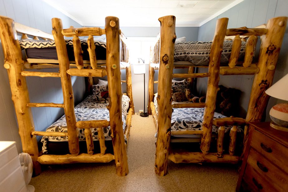 A cozy bedroom with two sets of rustic wooden bunk beds facing each other. The beds have patterned bedding, and there's a window in the center of the back wall letting in light. A small lamp sits on a wooden nightstand to the right.