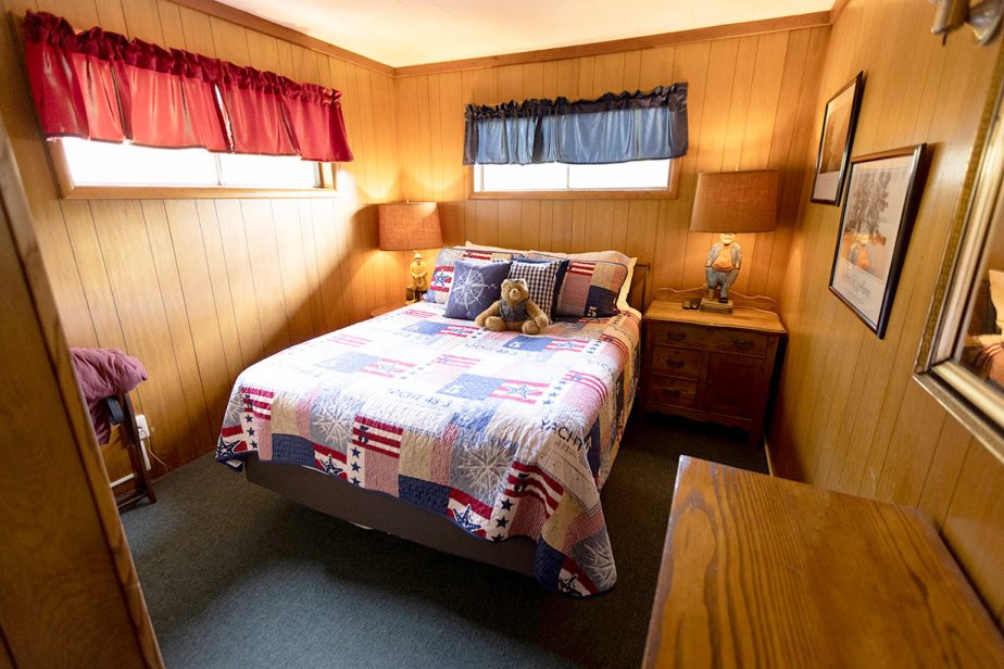 A cozy, wood-paneled bedroom with a large bed covered in a patriotic-themed quilt. Two side tables each host a bedside lamp, and a teddy bear sits on the bed among accent pillows. Red and blue valances decorate the top of the small windows, adding a charming touch.