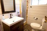 A modern bathroom with a rustic touch features a dark wood vanity with a white countertop and bronze fixtures under a framed mirror. A toilet is opposite the vanity, with a rustic door leading to a shower. Shiplap walls and wood-like flooring complete the look.