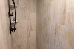 A modern shower with beige marble tile walls and hexagonal tile flooring. The shower features a rainfall showerhead on the ceiling and a handheld showerhead mounted on a rail. Toiletries are visible on built-in shelves in the corner.