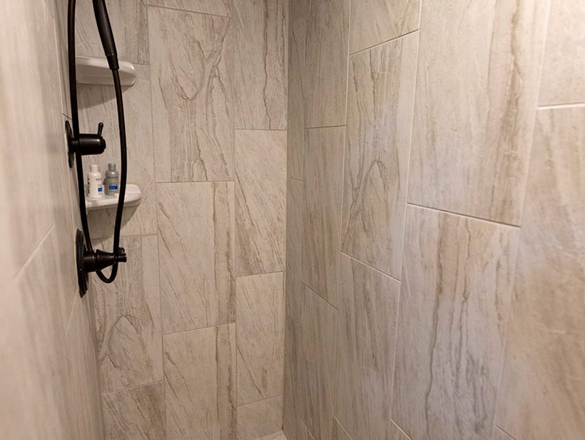 A modern shower with beige marble tile walls and hexagonal tile flooring. The shower features a rainfall showerhead on the ceiling and a handheld showerhead mounted on a rail. Toiletries are visible on built-in shelves in the corner.