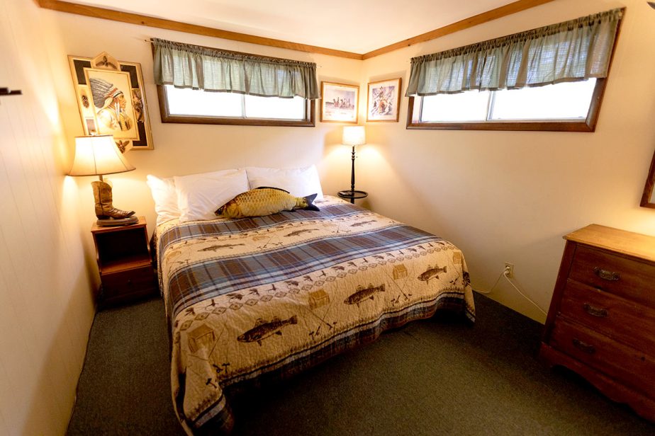 A cozy bedroom with a queen-sized bed covered in a fisherman's themed quilt. The room is lit by two decorative table lamps on wooden nightstands, with framed pictures of nature on the walls and two windows with green curtains allowing natural light in.