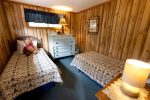 A cozy bedroom with wooden walls features two single beds adorned with patterned quilts and a teddy bear on one bed. A white dresser stands under a small window with a blue curtain, and a framed picture hangs on the wall. A lit lamp on a bedside table adds warmth.