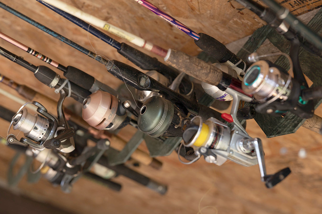 A close-up view of several fishing rods with reels mounted on a rack. The rods and reels vary in color and style, and the rack is mounted on what appears to be a wooden wall or ceiling. The focus is on the reels, showcasing their different designs.