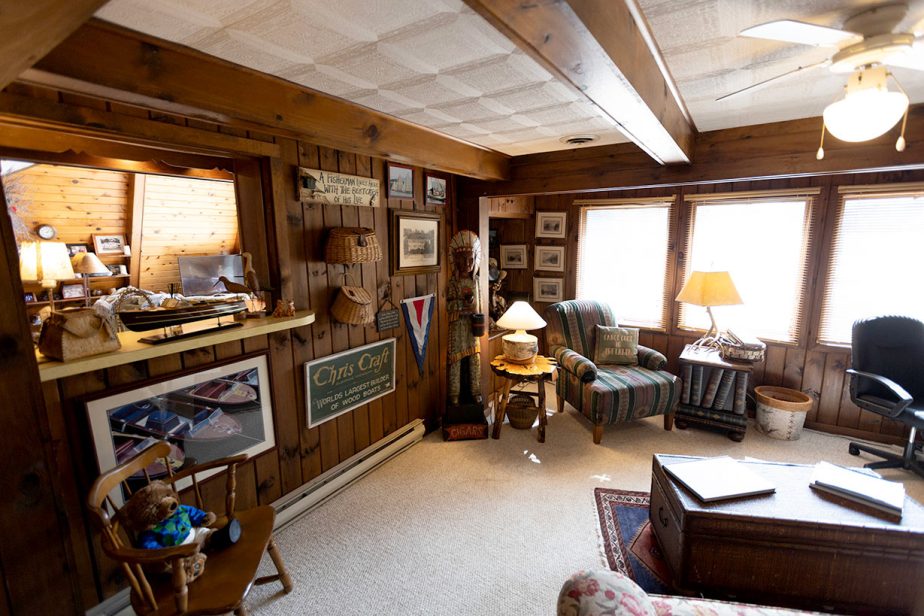 A cozy home office with wooden walls and large windows. The room features a desk, an office chair, an armchair with a side table, and various decorations, including framed photos and knick-knacks. Lamps provide warm lighting, and a ceiling fan is visible overhead.