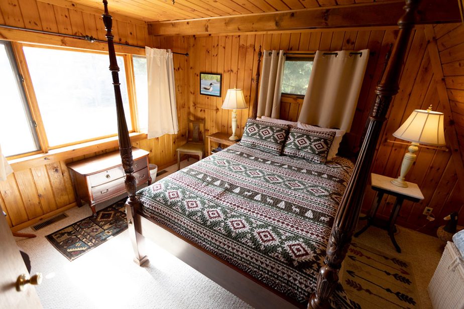 A cozy bedroom with wood-paneled walls features a large window with curtains, a four-poster bed with patterned bedding, two bedside tables with lamps, a wooden dresser, and a chair. A small painting hangs on the wall above the chair. Natural light fills the room.