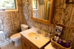 A small bathroom with patterned wallpaper resembling tree bark. It features a toilet, a wooden vanity with a white sink, and a lighted mirror. A window on the left wall provides natural light. A wicker basket on a small shelf holds toiletries.