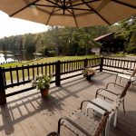 A spacious wooden deck with several chairs and potted plants overlooks a serene lake surrounded by trees. A large umbrella provides shade over part of the deck, and in the background, there is a large building with more seating and a boat dock by the water.