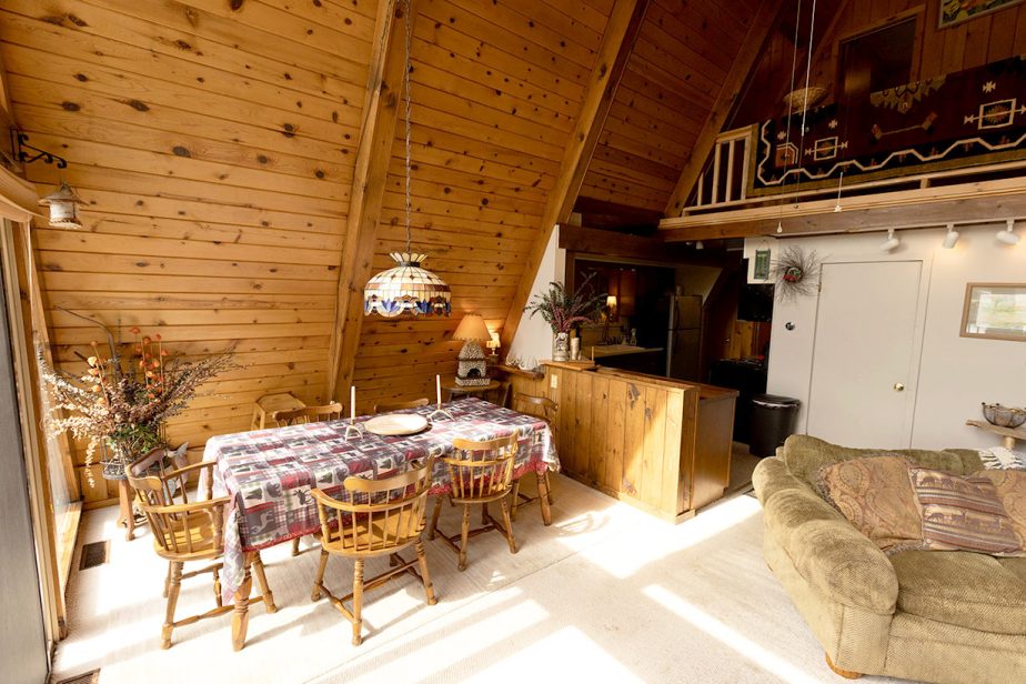 A cozy A-frame cabin interior with wooden walls and ceiling, showcasing a dining area with a table and six chairs covered in a patterned tablecloth. A beige couch sits nearby, and a kitchenette features a breakfast bar. Stairs lead to a small loft area.