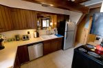 A cozy kitchen with wooden cabinetry and beige countertops. It features a stainless steel refrigerator, dishwasher, and electric stove. The sink area is illuminated by a small window, and various kitchen items are neatly arranged on the counters. The floor is tiled.