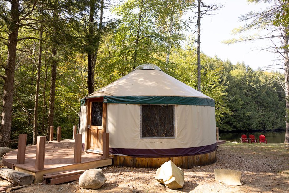 A yurt stands in a wooded area with a small deck on its side. The structure has a beige canvas with green trim, wooden door, and clear windows. In the background, two red chairs sit by a peaceful pond surrounded by tall trees.