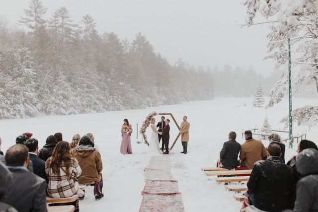 A wedding ceremony in a snowy outdoor setting. The bride and groom stand under an arched floral arrangement, accompanied by officiant and bridesmaid. Guests, dressed in winter clothing, sit on benches covered in snow, surrounded by snow-covered trees and landscape.