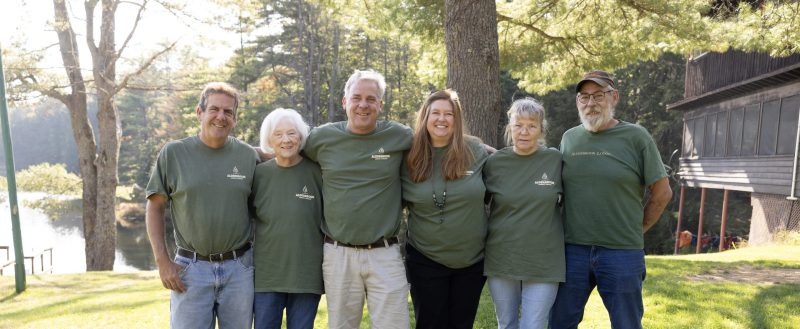 A group of six people wearing matching green shirts stand outdoors on a grassy area. They are smiling and have their arms around each other’s shoulders. Trees and a body of water are visible in the background. The setting appears to be a sunny day.