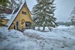 A cozy, yellow A-frame cabin is nestled among tall pine trees, surrounded by a thick blanket of snow. Footprints lead to the front door, which is adorned with a wreath. A wooden deck wraps around the side of the cabin, and a wintry landscape stretches out in the background.