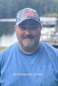 A man wearing a blue T-shirt with "Alderbrook Lodge" written on it and a camouflage cap featuring the American flag stands outdoors near a wooden dock and a calm lake with a wooded area in the background. The atmosphere appears serene and peaceful.